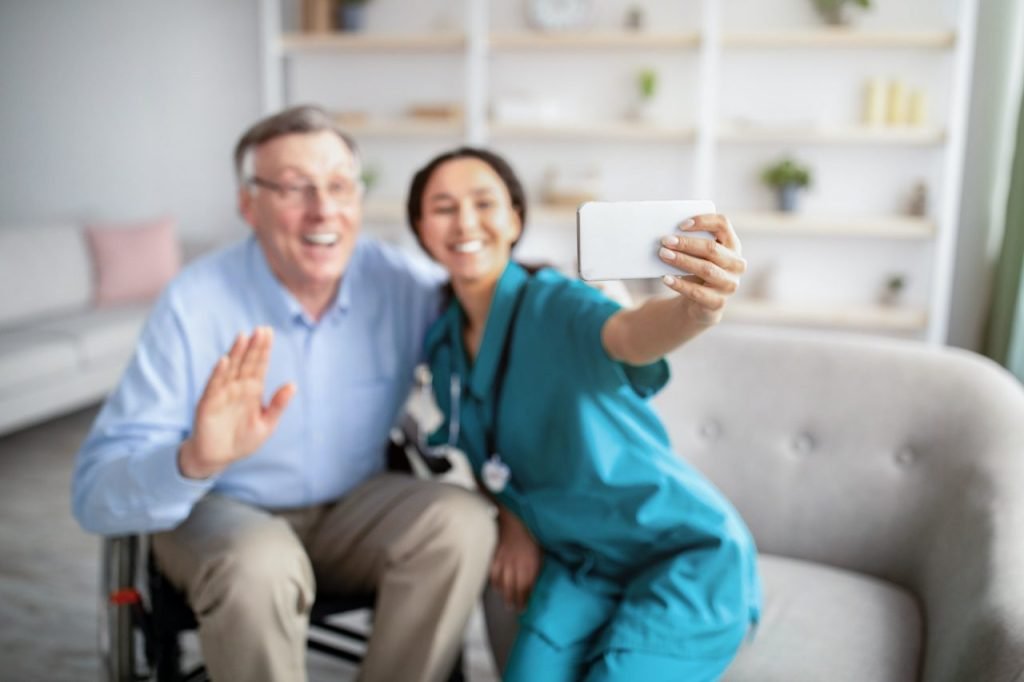 cheerful disabled senior man in wheelchair and his nurse taking selfie indoors selective focus
